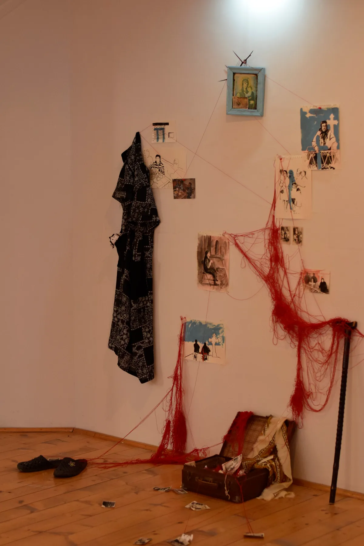 Full installation view of Mamaia Vasilica with Orthodox icon at the apex, drawings, robe, walking stick, slippers, and open suitcase on the floor