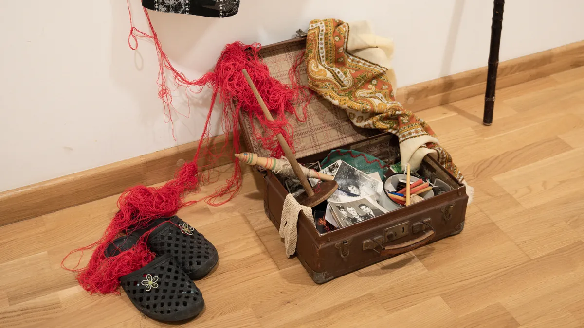 Floor-level installation objects: slippers, open suitcase, wooden distaff, red wool thread, and walking stick on the wooden floor